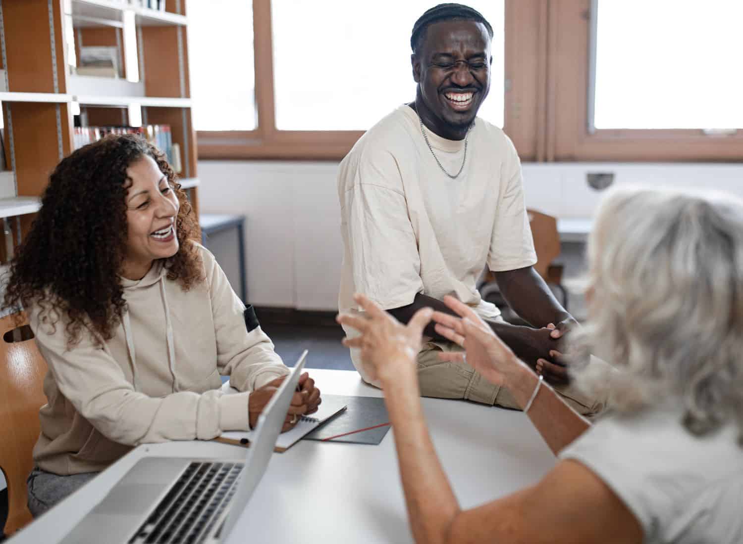 Three people sitting at a table in a library, laughing together. Two younger individuals, a man and a woman, are facing a smiling older woman. A laptop with notes on New York Bartending Classes and notebooks are on the table, while bookshelves stand proudly in the background.