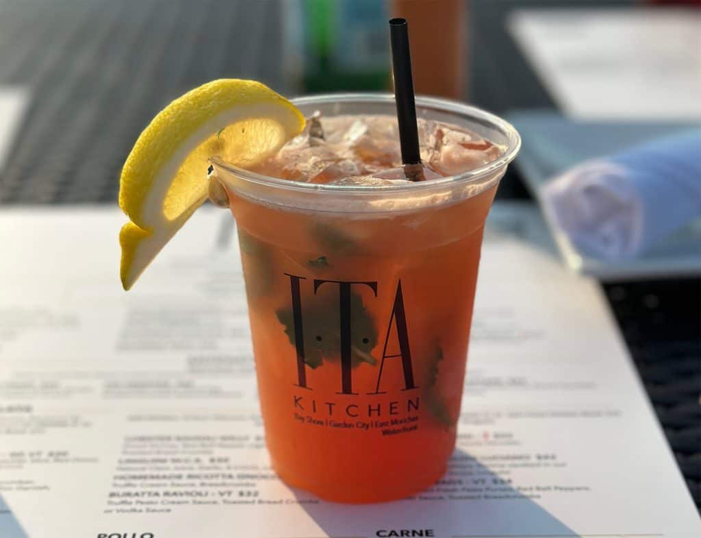 A plastic cup filled with an iced beverage, garnished with a lemon wedge and a black straw, evokes the flair learned at Queens Bartending Classes. The cup is labeled "ITA Kitchen." A blurred menu is visible in the background on an outdoor table.