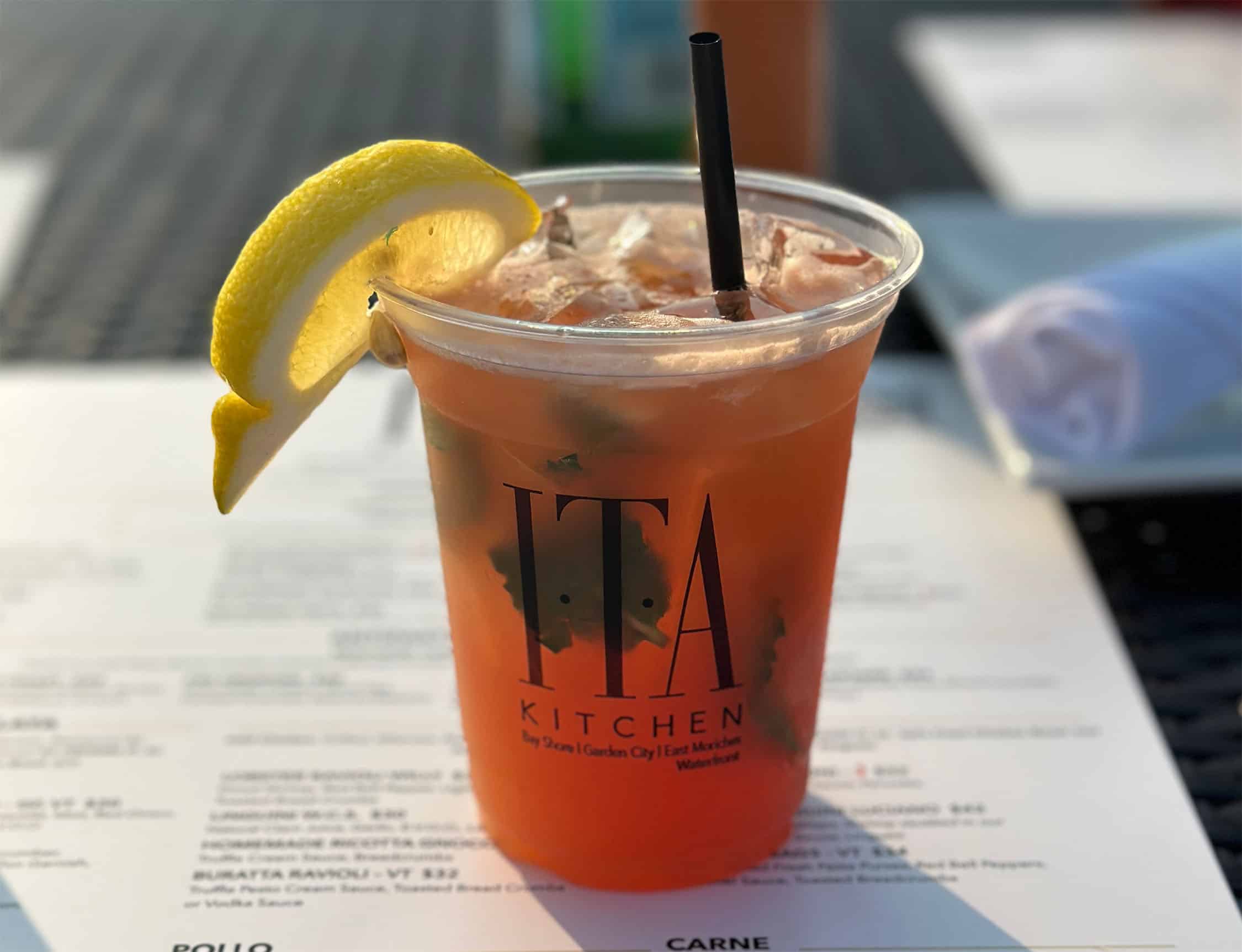 A plastic cup filled with an iced beverage, garnished with a lemon wedge and a black straw, evokes the flair learned at Queens Bartending Classes. The cup is labeled "ITA Kitchen." A blurred menu is visible in the background on an outdoor table.