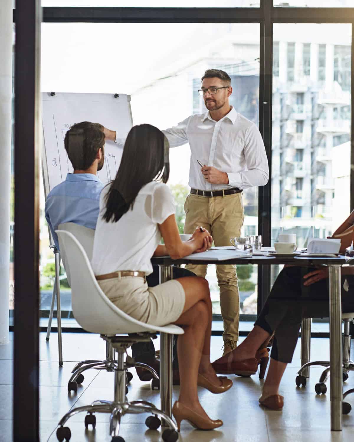 A man in a white shirt is presenting at a whiteboard to three colleagues sitting at a conference table. The room, reminiscent of the sleek spaces used for Long Island Bartending Classes, boasts large windows with a city view. The group appears focused and engaged.