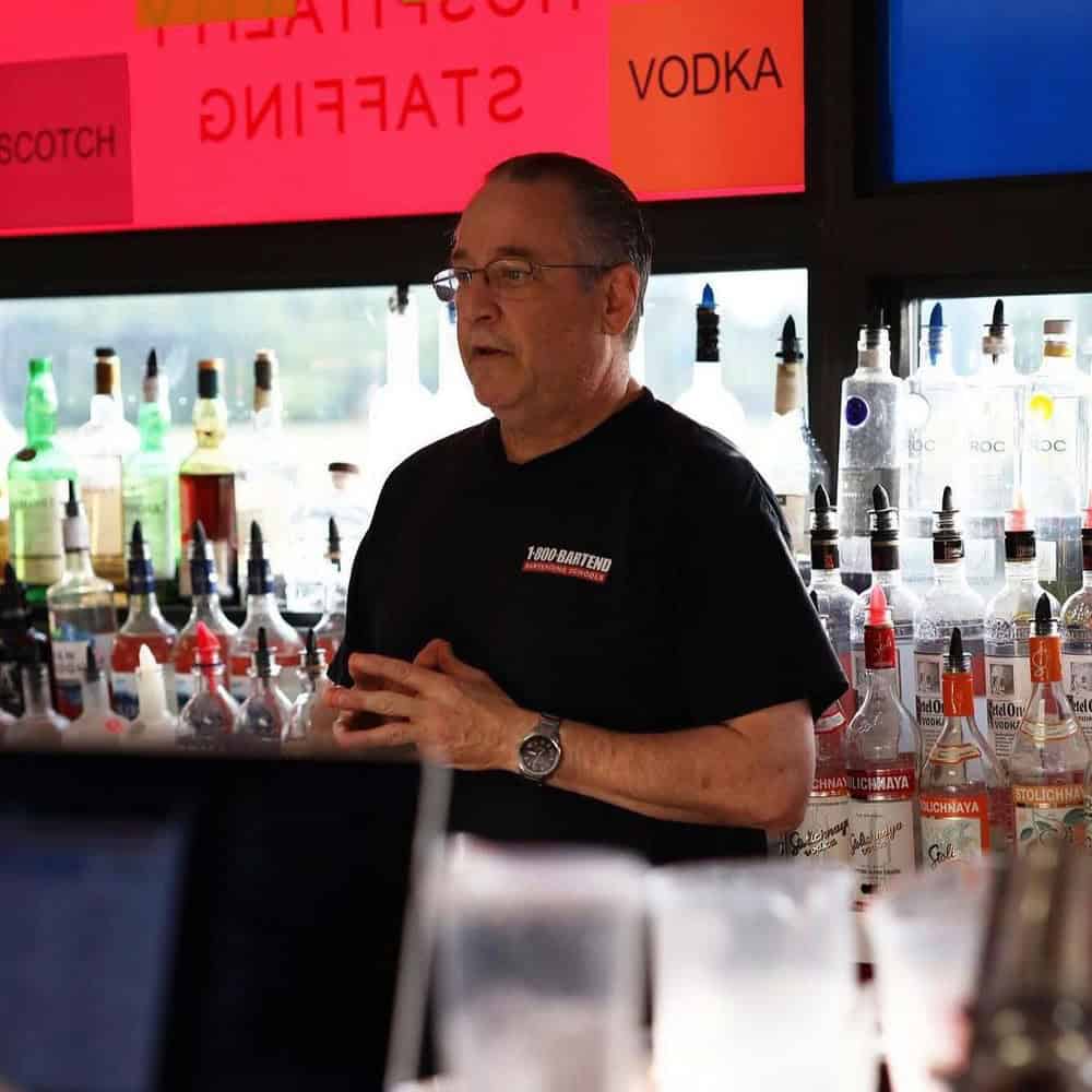 A man stands behind a bar counter stocked with various bottles of liquor, perhaps during a session at the Long Island Bartending School. He is speaking or giving a demonstration. Brightly colored signs are visible in the background, indicating different types of alcohol like vodka and scotch.