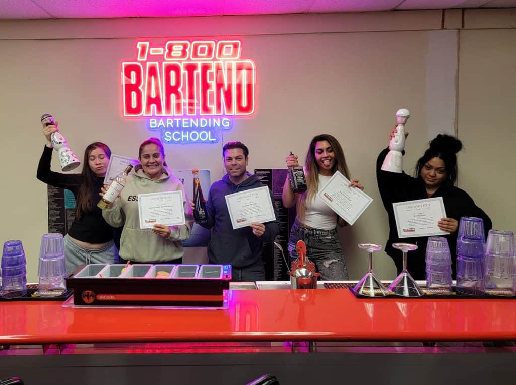 Five people stand behind a bar counter, each holding certificates and bottles. A neon sign above reads "1-800 BARTEND BARTENDING SCHOOL." Various bar tools are on the counter in the foreground—a proud moment at Queens Bartending School.
