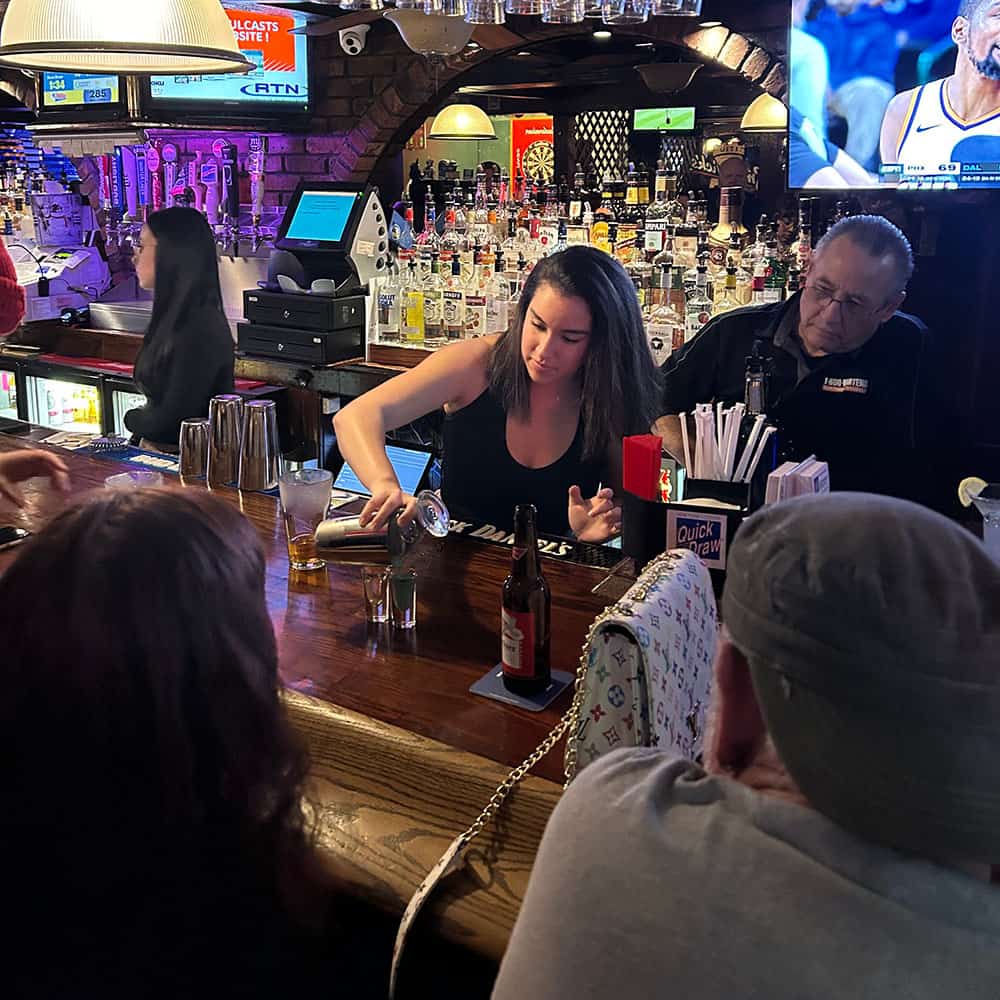 A bartender, trained at New York Bartending School, pours a drink at a bustling bar with patrons seated at the counter. A TV screen in the background shows a sports game, and the shelves are stocked with various bottles.