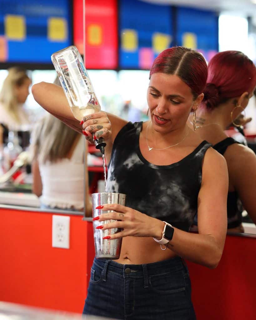 A bartender with red hair, wearing a black and white crop top, pours a clear liquid from a bottle into a metal shaker. She deftly mixes cocktails at the brightly-colored bar, showcasing skills honed in Queens Bartending Classes. Mirrored walls and an open window frame the vibrant scene.