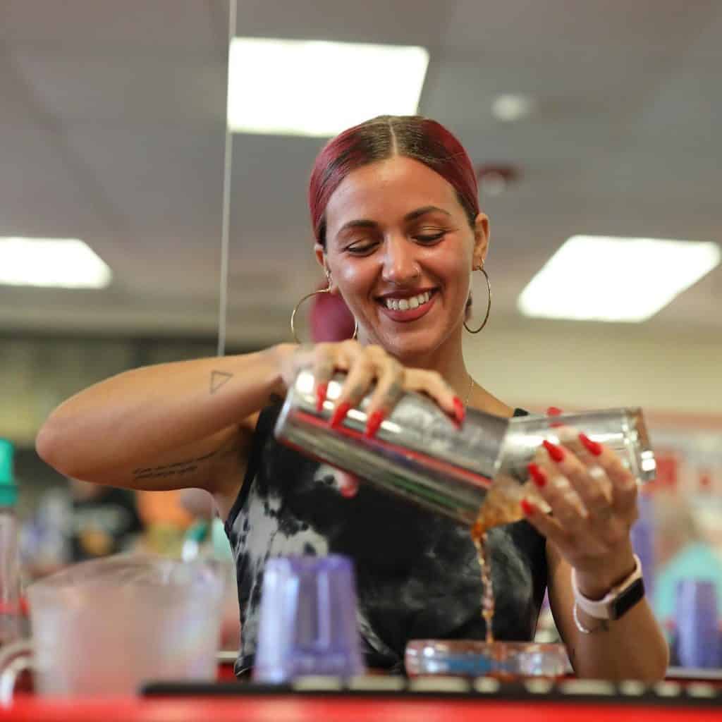 A person with red hair smiles while pouring a drink from a cocktail shaker into a glass, showcasing their skills from Long Island Bartending Classes. Wearing hoop earrings and a sleeveless top, they are surrounded by various bar tools and bottles.