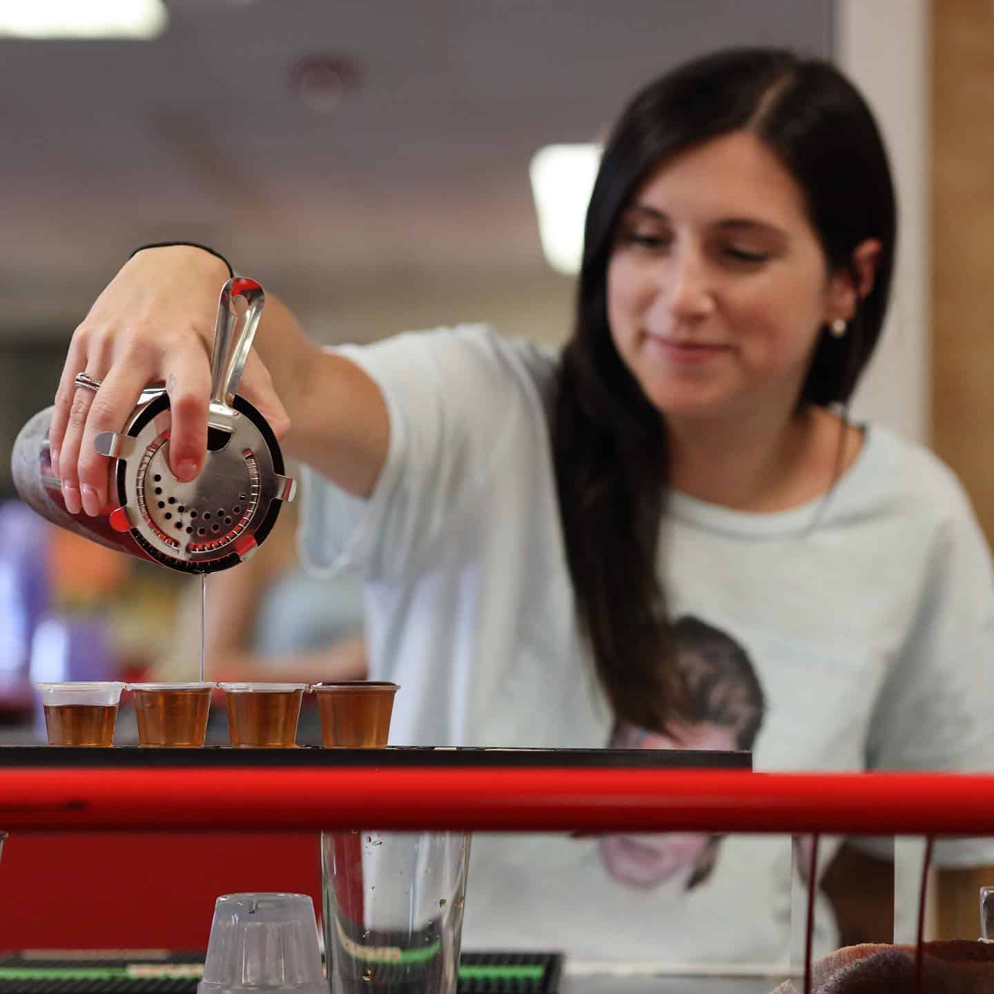 A woman, perhaps a graduate of New York Bartending School, is pouring drinks from a cocktail shaker into plastic cups on the bar counter. Clad in a light-colored shirt and smiling softly, she expertly uses various bartending tools as she focuses on the pour.