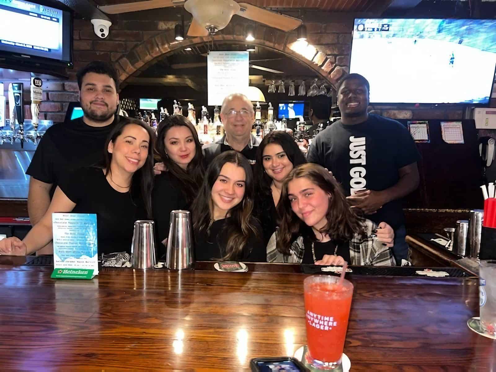 A group of people smiling and posing behind a bar counter, showcasing their skills from New York Bartending Classes. Drinks and bar equipment are in the foreground, with TV screens adding ambiance to this lively bar or restaurant setting.