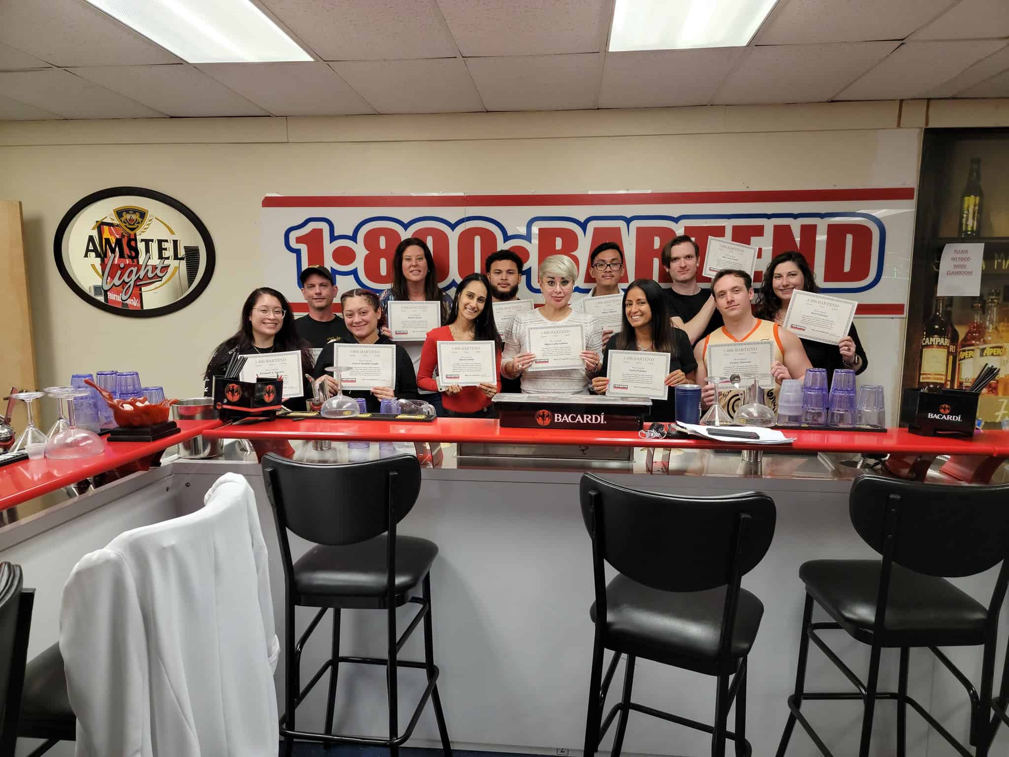 A group of people stand behind a bar, each holding a certificate from Long Island Bartending School. The setting includes bar stools, alcohol bottles, and signs for Amstel Light and Bacardi. They are smiling and appear to be celebrating a shared achievement.