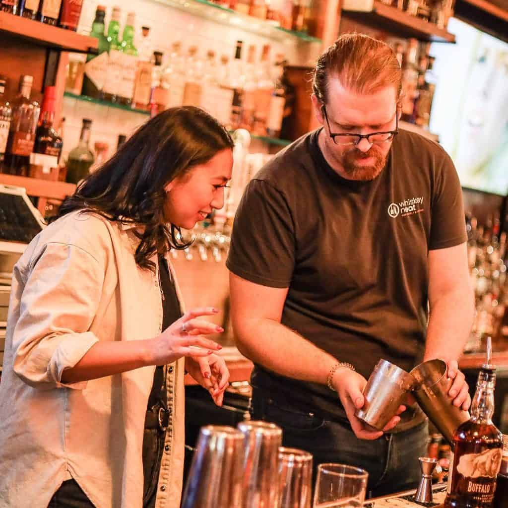 A man and a woman stand behind a bar counter at New York Bartending School. The man, wearing a black T-shirt, pours a drink into a shaker. The woman smiles and gestures with interest. Shelves filled with bottles are visible in the background.