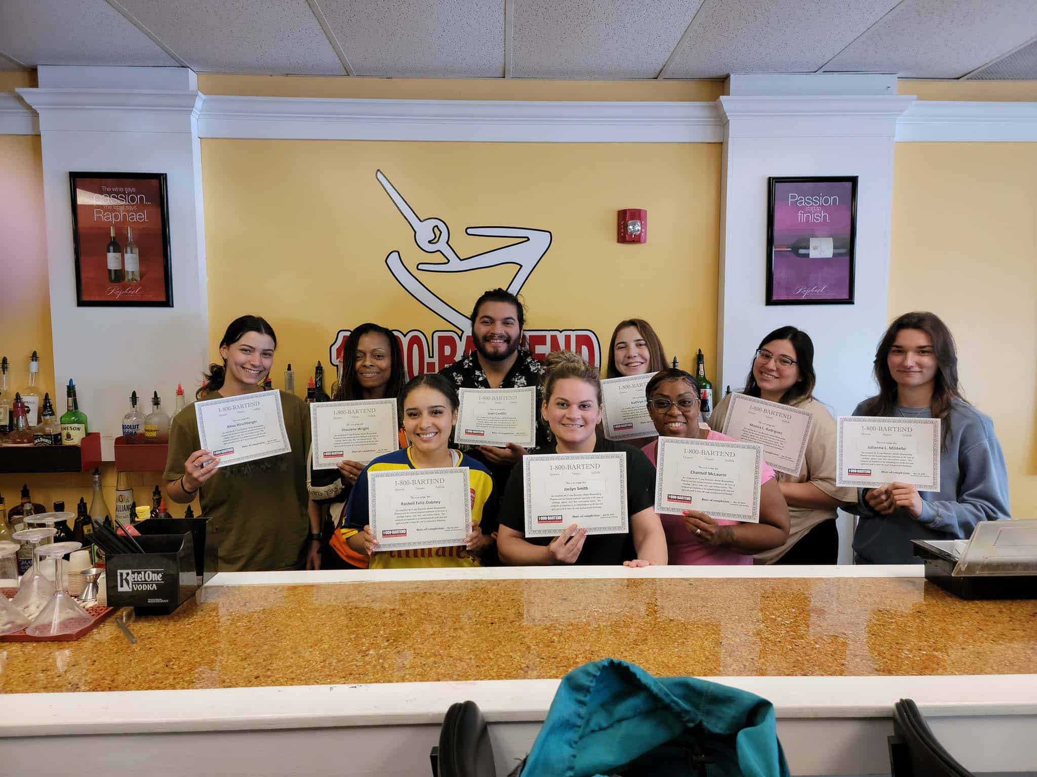 A group of nine people stands behind a bar, each proudly holding a certificate from the Queens Bartending School. They are smiling and posing for the camera against a yellow wall adorned with the "School of Bartending" logo and two posters, with bottles elegantly displayed on shelves.