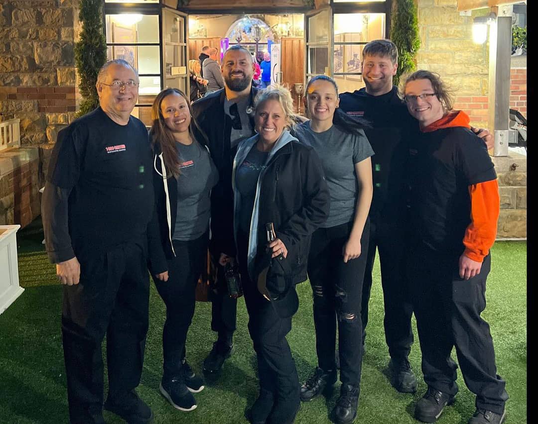 A group of seven people are standing together outdoors, smiling at the camera. Dressed in casual matching attire, they pose in front of a building with an open door. The setting appears to be in the evening, perhaps after a session at Long Island Bartending School.