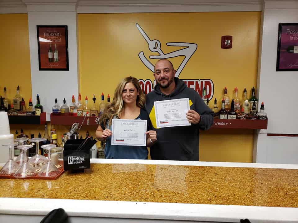 Two people stand behind a bar counter holding certificates from Queens Bartending School. The background features shelves with various liquor bottles and a logo with a martini glass. The man is wearing a hoodie, and the woman is in a long-sleeve top.