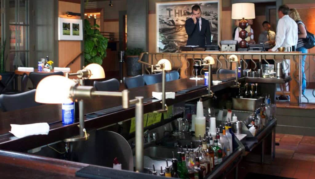 A restaurant bar with an assortment of bottles and lamps showcases its ambiance. In the background, a man in a suit uses a phone while a painting hangs on the wall. Several people are near the entrance, perhaps discussing their latest tips learned at New York Bartending Classes.