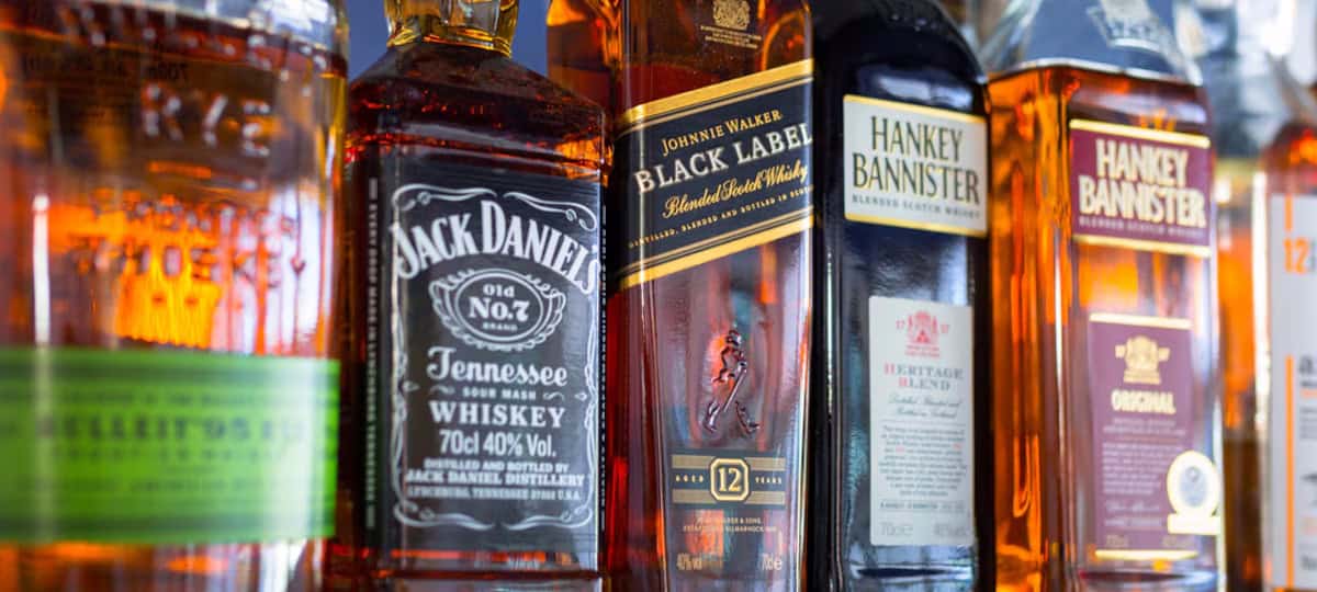 Bottles of whiskey and other spirits, featuring well-known brands with distinct labels, line the shelf in vibrant colors and designs. This diverse collection mirrors the expert training at Long Island Bartending Classes, where creative mixology comes to life.