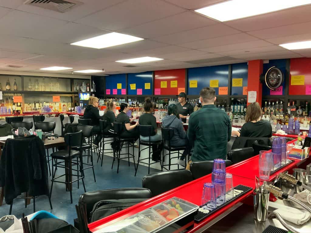 A group of people sits on bar stools in a brightly lit bar in Queens, its red, blue, and yellow walls echoing the vibrant energy of nearby Bartending School. Various bottles line the shelves behind the bar, adorned with colorful sticky notes. Glasses and tools clutter the counter.
