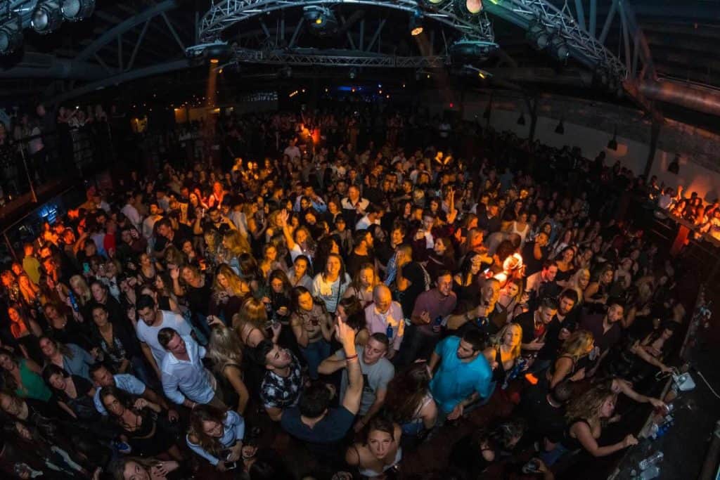A large crowd of people enjoying a lively indoor concert or party in New York. The image shows a diverse group of individuals, possibly including some from local bartending classes, dancing and socializing under colorful stage lights in a spacious venue with visible metal beams and a balcony area.