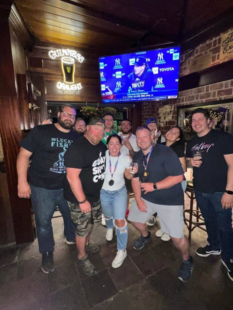 A group of people smiling and posing together in a bar with a sports game on TV in the background, possibly celebrating after a fun class at Queens Bartending School. Some are holding drinks, and one person is proudly wearing a medal. The atmosphere appears lively and cheerful.