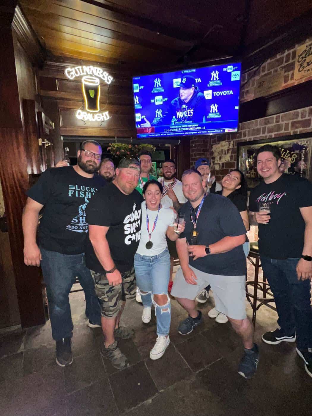 A group of people smiling and posing together in a bar with a sports game on TV in the background, possibly celebrating after a fun class at Queens Bartending School. Some are holding drinks, and one person is proudly wearing a medal. The atmosphere appears lively and cheerful.