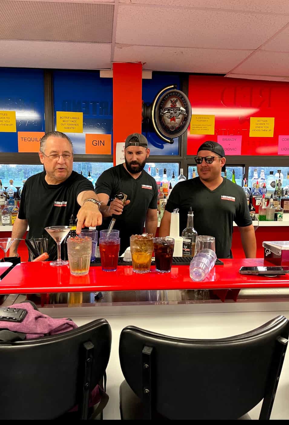 Three men stand behind a red bar counter filled with assorted drinks and bottles. They're dressed in black shirts, and one is pouring a drink. Various spirits are visible in the background, reminiscent of a practice session at Queens Bartending School. The atmosphere appears friendly and casual.