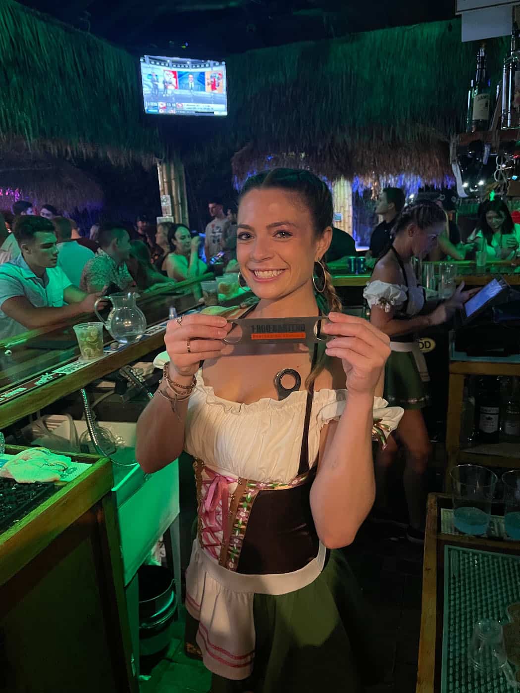 A woman in traditional attire stands in a lively bar, smiling and holding a card with "Bong Barriga" written on it. The festive atmosphere might remind one of graduation nights at the New York Bartending School. The bar teems with people, while a TV flickers in the background.