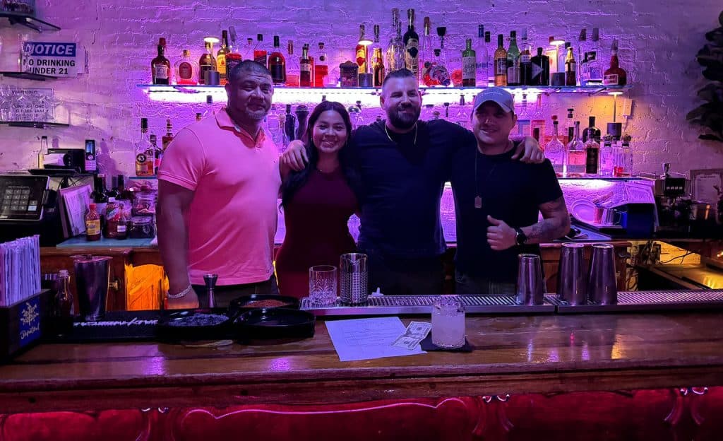 Four people stand together behind a dimly lit bar, smiling. On the counter in front of them are various beverages and cocktail-making tools, showcasing their skills from Queens Bartending Classes. The bar shelves behind them are lined with illuminated bottles.