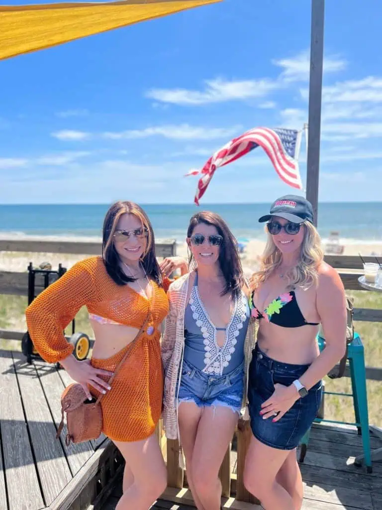 Three women in beach attire stand on a wooden deck with an ocean view, smiling at the camera. An American flag waves proudly in the background, evoking a sense of patriotism. It's a perfect summer day—a stark contrast to their fun-filled evenings at New York Bartending School.