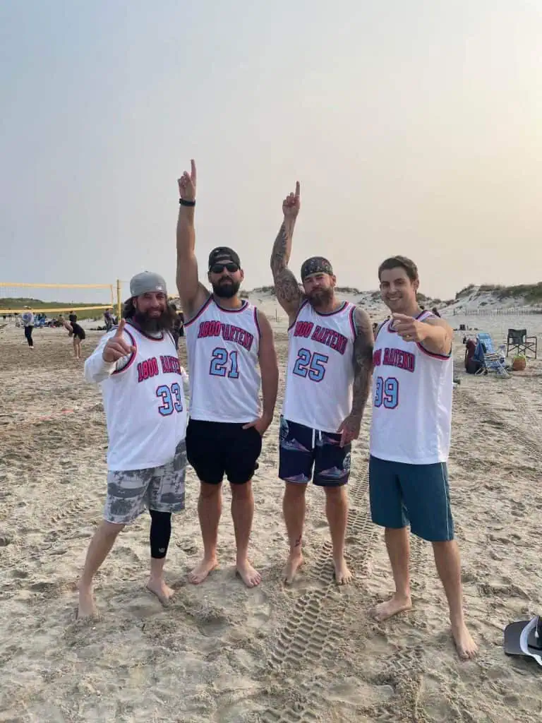 Four men stand together on a sandy beach, wearing matching white jerseys with red and blue numbers. They're pointing upwards with one hand, smiling, and a volleyball net is visible in the background, perhaps celebrating a win after attending New York Bartending School's fun team-building event.
