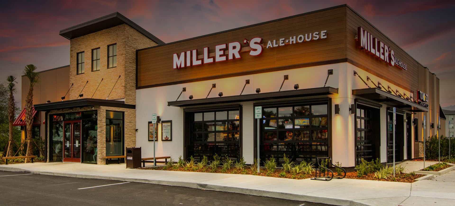 Miller's Ale House, with its modern facade and outdoor lighting at dusk, offers a warm welcome to those strolling by. Located near the Queens Bartending School, the restaurant features large windows and a mix of brick and wood paneling, surrounded by lush greenery and a paved sidewalk.