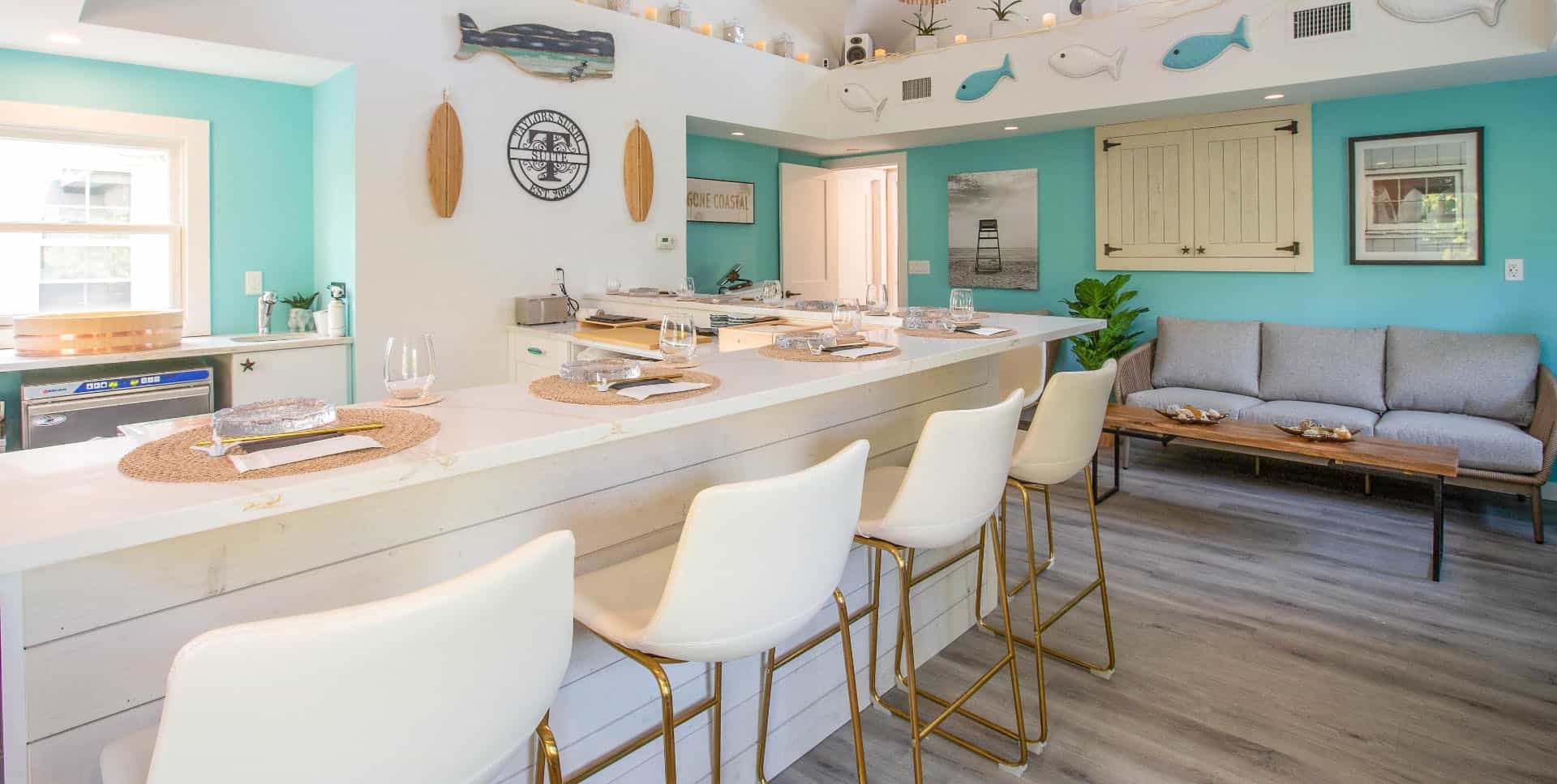 A bright kitchen with teal walls features a long white island reminiscent of a stylish bar scene, complete with nautical decorations and beach-themed decor. It's as inviting as the atmosphere at Long Island Bartending School, with a cozy seating area to the right, adorned by potted plants and artworks.