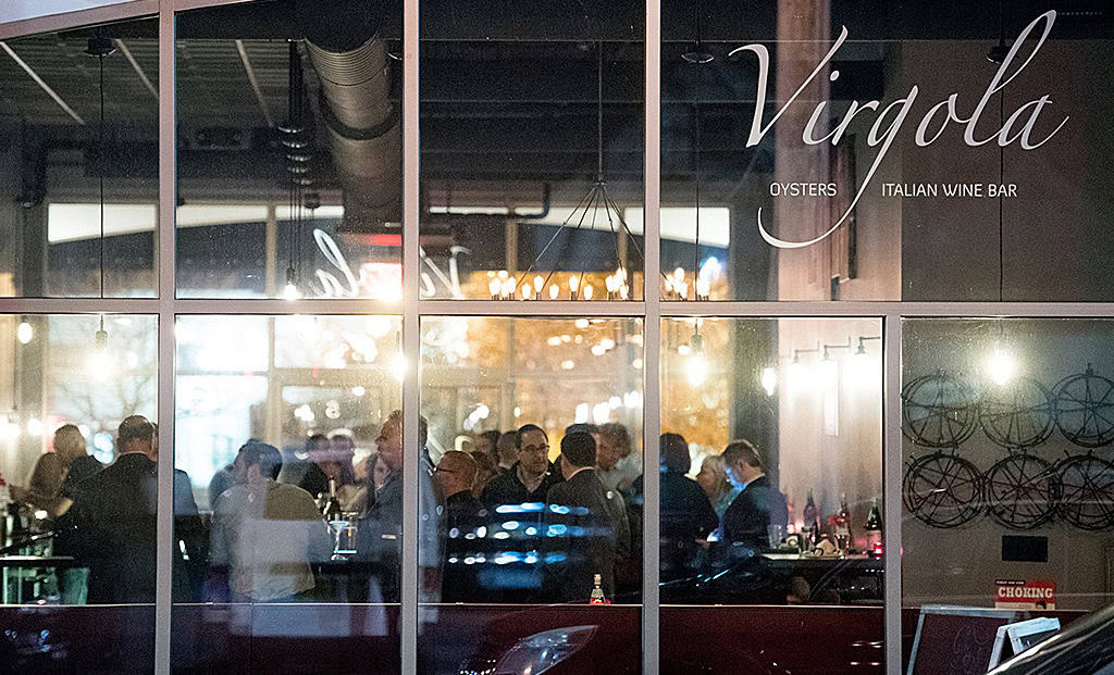 View through the window of a bustling Italian wine bar, Virgolao, filled with people socializing. Warm lighting and modern decor create a cozy ambiance, reminiscent of lively gatherings at New York Bartending School events. A car is partially visible outside.