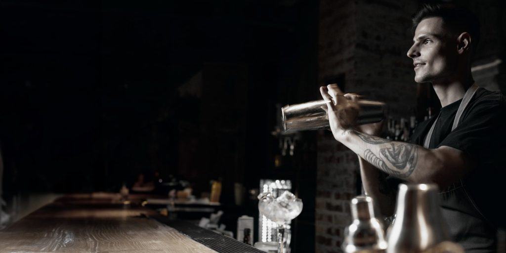 A bartender with tattoos, a graduate of New York Bartending School, is shaking a cocktail shaker behind a dimly lit bar. A glass with ice sits on the bar counter. The atmosphere is moody and focused.