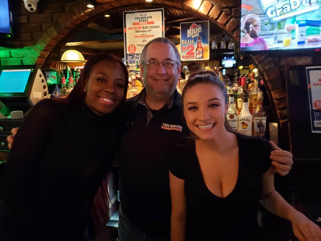 Three adults smiling in a bar setting, perhaps graduates from Queens Bartending School. Two women flank a man, standing in front of a well-stocked bar. A TV screen and various bottles are visible in the background, creating a lively atmosphere.