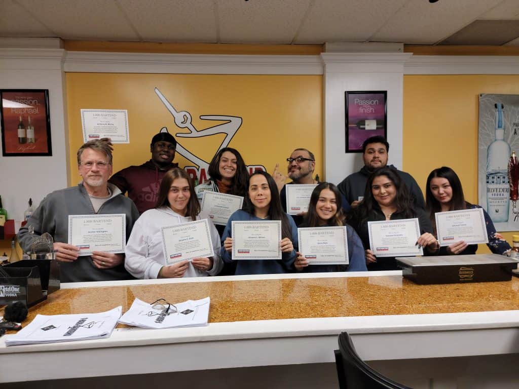 A group of ten people smiling and holding certificates, standing in a classroom with a yellow wall and motivational posters. Fresh from Queens Bartending School, they've just completed their course. A table with papers is in front of them, capturing their success in mixology mastery.
