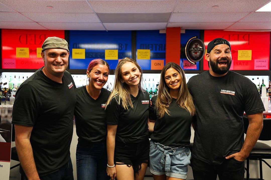 Five people are smiling and posing together inside a bar with various colorful signs in the background. Wearing black shirts, they stand closely side by side, perhaps celebrating their time at Queens Bartending Classes.