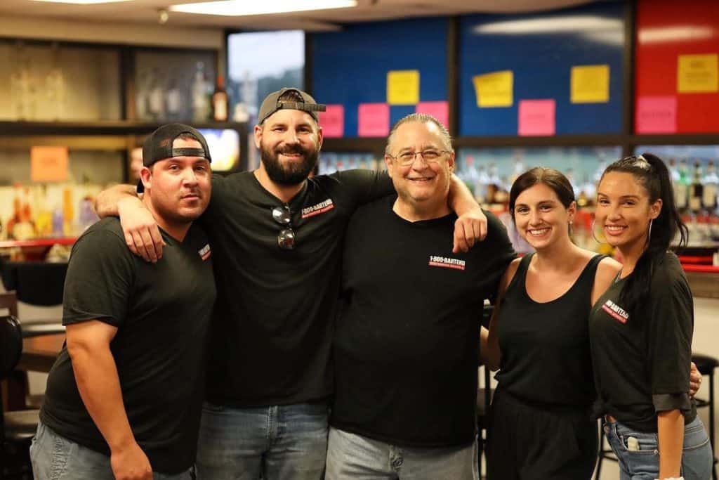 A group of five people, fresh from their New York Bartending Classes, stands closely together, smiling at the camera in a casual indoor setting. They all wear black shirts, and the background features shelves with various items and colorful notes on a wall.
