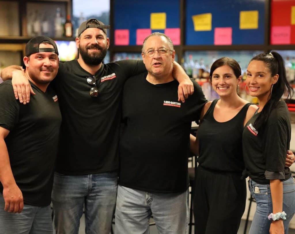 A group of five people in casual black shirts stand together indoors, smiling for the camera. They appear to be in a lively social setting or a restaurant, perhaps celebrating after a fun session at New York Bartending School. Bright colored menus or signs are visible in the background.