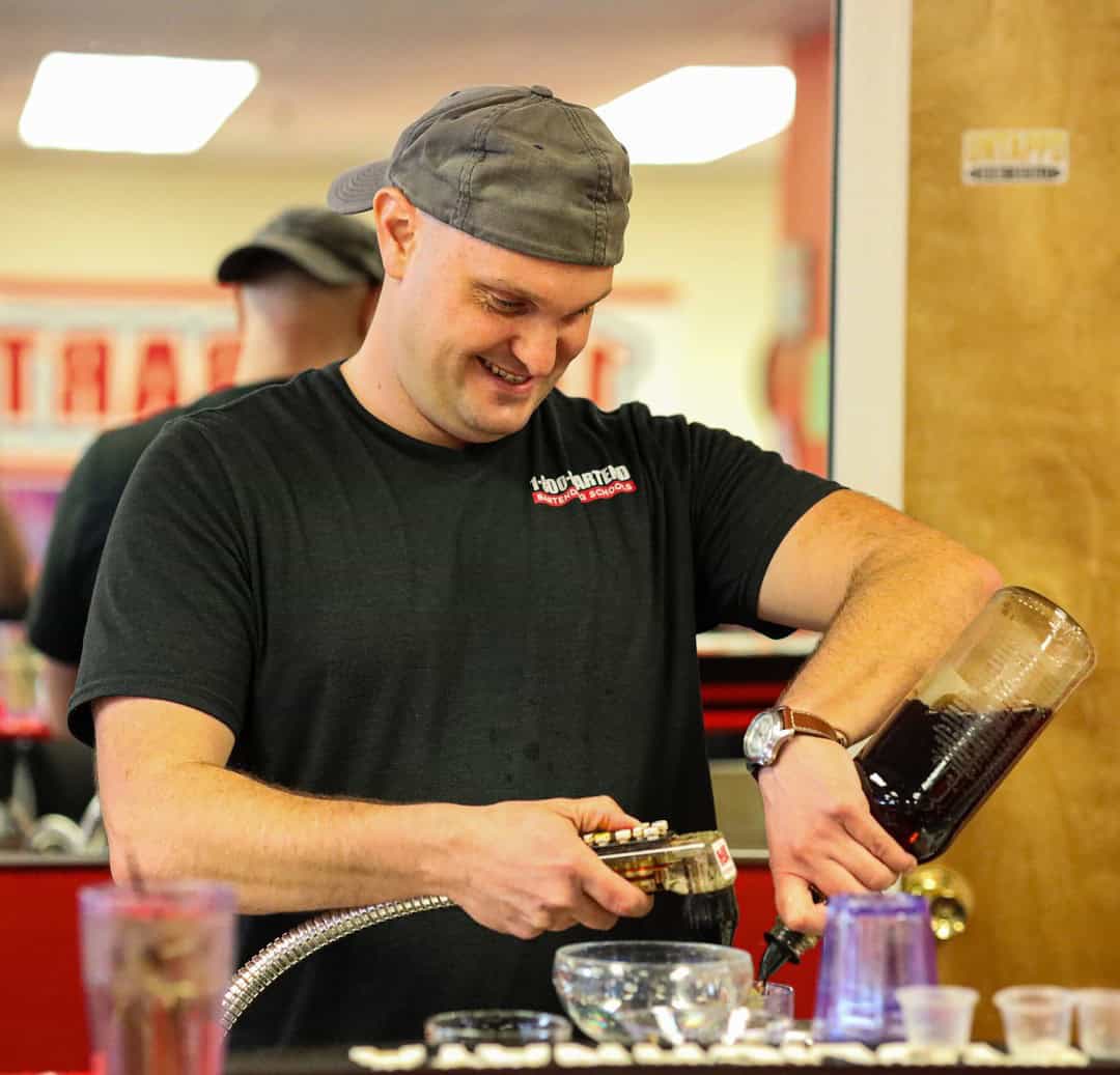 A man wearing a cap and a black T-shirt is smiling while bottling a dark liquid at a counter, perhaps showcasing his skills from the Long Island Bartending School. Clear cups and bottles surround him, creating a casual and lively atmosphere.