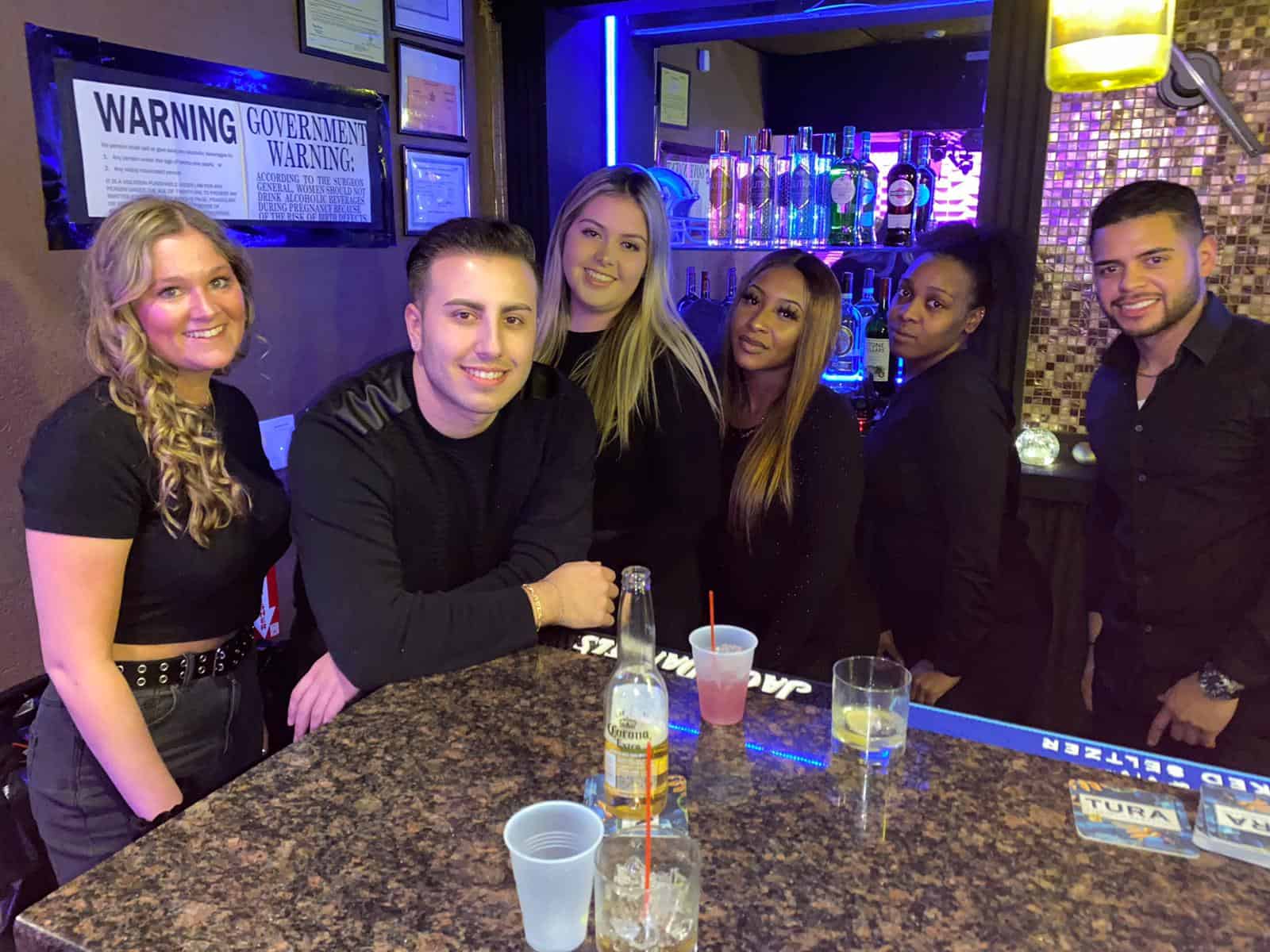A group of six people, three men and three women, stand behind a bar counter filled with drinks and bottles. They are all smiling, casually dressed in dark clothing. With a 'Government Warning' sign in the background, it looks like they've just completed their course at New York Bartending School.