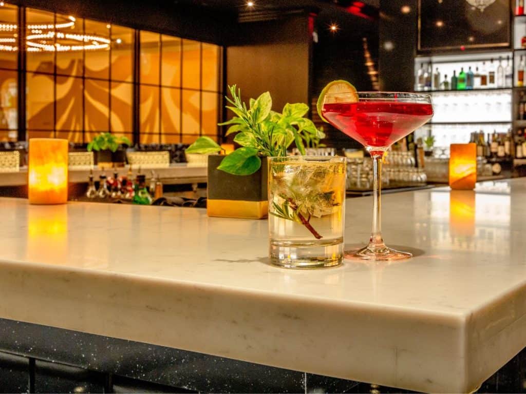A stylish bar setting features a glossy white marble counter. On the counter are two cocktails: a vibrant red martini garnished with a citrus slice and a clear drink with herbs. Warm lighting and plants add to the ambiance, reminiscent of what students create in Long Island Bartending Classes.