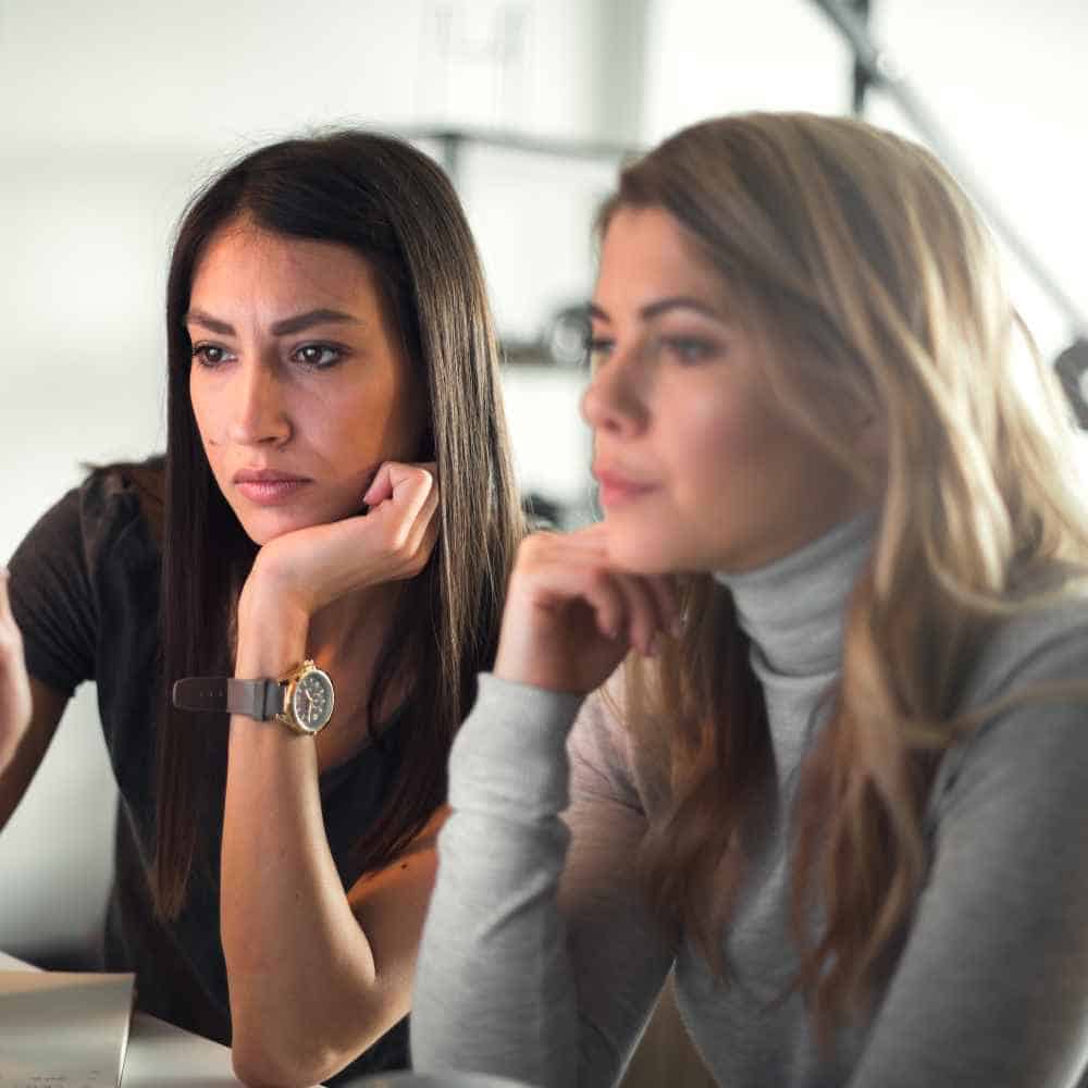 Two women sit together, intently focused on a computer screen. The woman on the left, with long dark hair and a watch, and the woman on the right, in a gray sweater with long light hair, both rest their chins on their hands. Perhaps they're exploring Bartending School NY online courses.