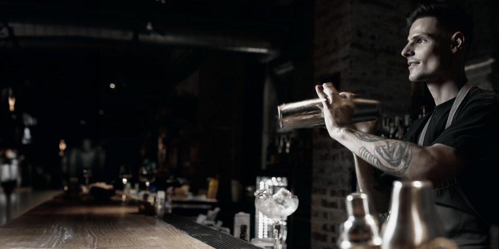A tattooed bartender, trained at Queens Bartending School, skillfully shakes a cocktail shaker in the dimly lit bar. The wooden counter is lined with various glasses and bottles, creating a focused and professional atmosphere.