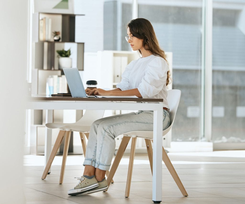 A woman with long hair and glasses sits at a white desk, typing on a laptop. Dressed in a white blouse and light jeans, she sips coffee while researching Bartending School NY. The room is bright with large windows and modern decor.