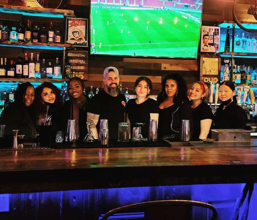 A group of seven people behind a bar counter pose for a photo in a dimly lit venue. Shelves of bottles and a TV displaying a soccer match set the scene, perhaps capturing graduates from Queens Bartending Classes celebrating their skills with newfound camaraderie.