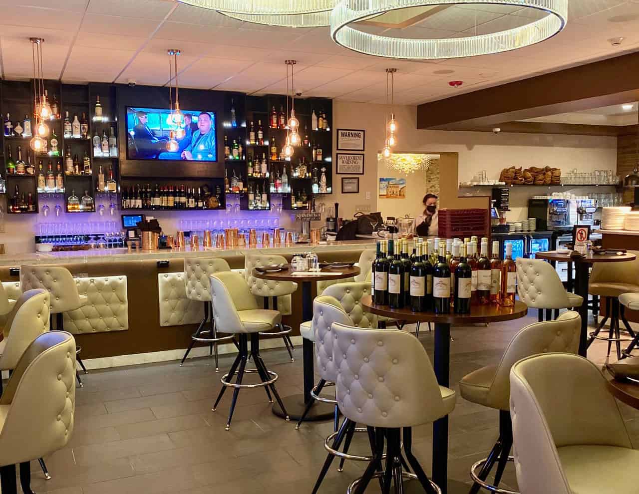 A cozy bar and restaurant interior featuring cushioned chairs, a variety of wines on display, and a wall-mounted TV under dim lighting. The bar in the background is reminiscent of the fine techniques taught at Queens Bartending School, with shelves stocked with bottles and glasses.