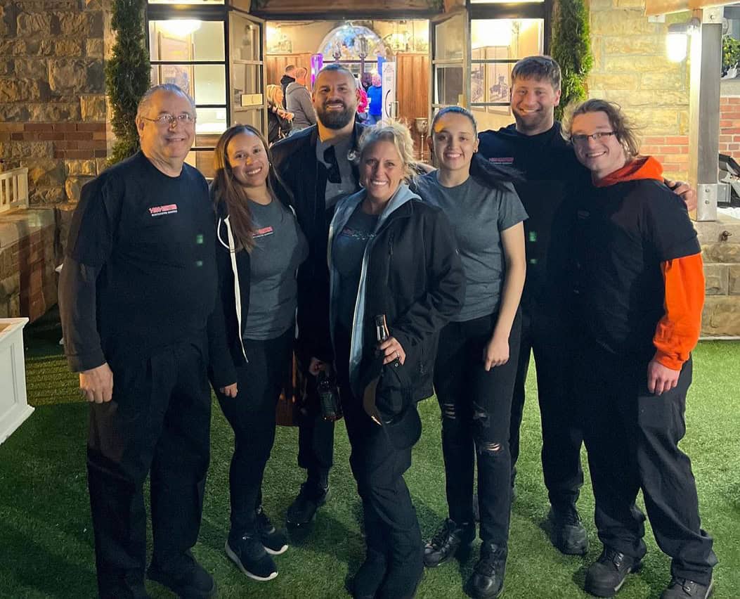 A group of seven people, dressed in casual attire, pose for a photo outside the Long Island Bartending School during the evening. The background shows an open doorway with people inside. Everyone is smiling.