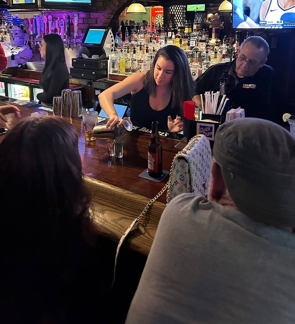 A female bartender with long hair, trained at Queens Bartending School, pours a drink at a dimly lit bar. Two customers sit in front, engaged in conversation. Bottles are visible in the background, and a man sits next to the bartender. A TV screen is on above.