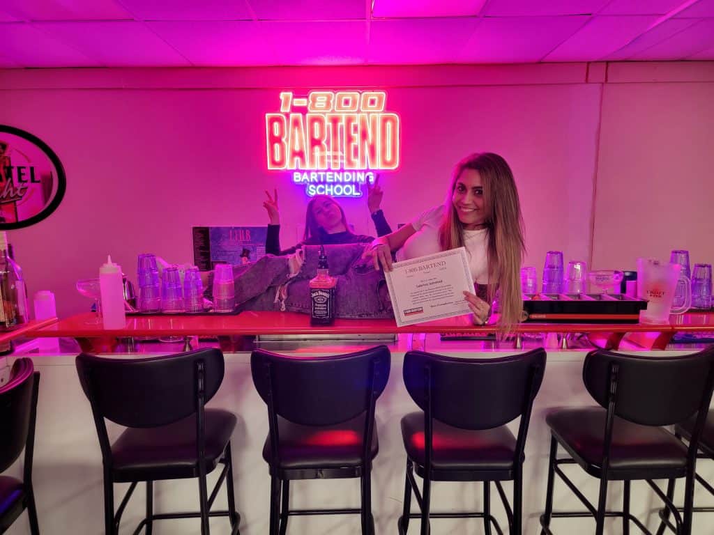Two people stand behind a bar, one holding a certificate and smiling at the camera. A neon sign above reads "1-800 Bartend Bartending School." The bar boasts various bottles and glasses, with four stools in front. This scene is a testament to the top training at Long Island Bartending School.