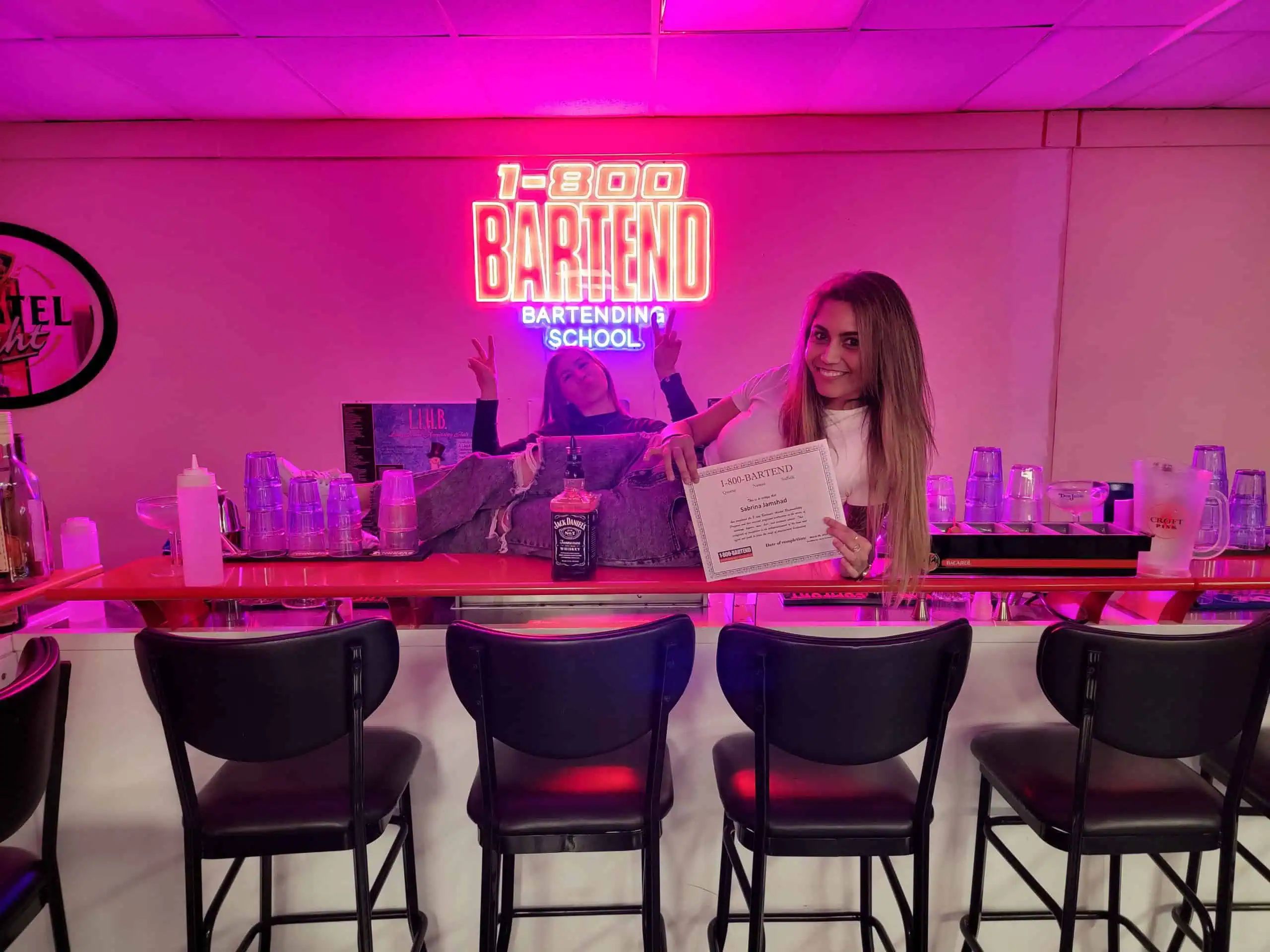Two people stand behind a bar, one holding a certificate and smiling at the camera. A neon sign above reads "1-800 Bartend Bartending School." The bar boasts various bottles and glasses, with four stools in front. This scene is a testament to the top training at Long Island Bartending School.