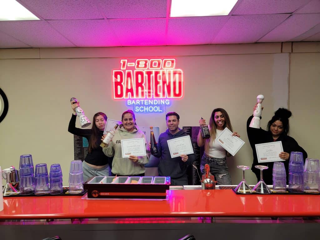 Five people posing at a bar counter, each holding a bartending tool. Three proudly display certificates from New York Bartending School. A neon sign above them reads "1-800-BARTEND Bartending School." The counter glistens with cocktail shakers and glasses, capturing the essence of skilled mixology.