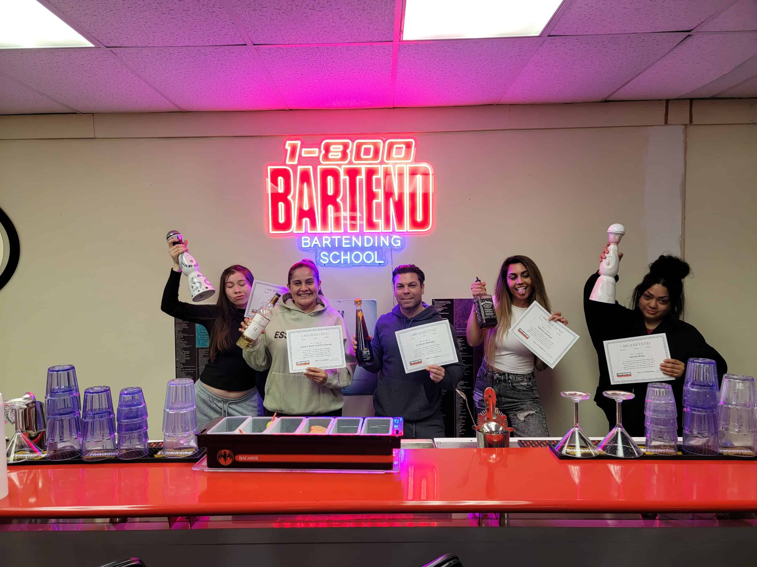 Five people posing at a bar counter, each holding a bartending tool. Three proudly display certificates from New York Bartending School. A neon sign above them reads "1-800-BARTEND Bartending School." The counter glistens with cocktail shakers and glasses, capturing the essence of skilled mixology.